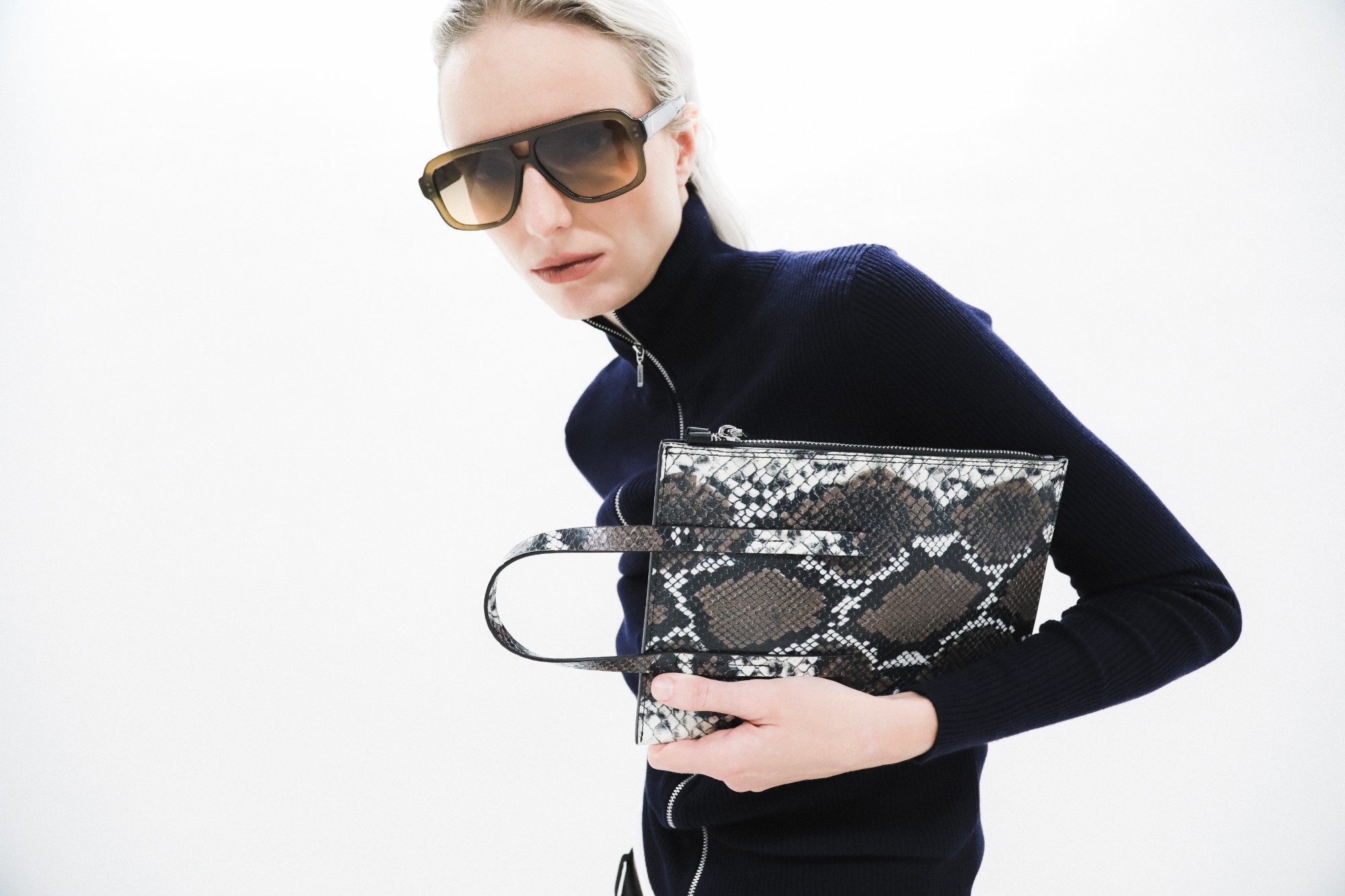 Model in a navy zip-front sweater holding a snake-print DIENÈE clutch